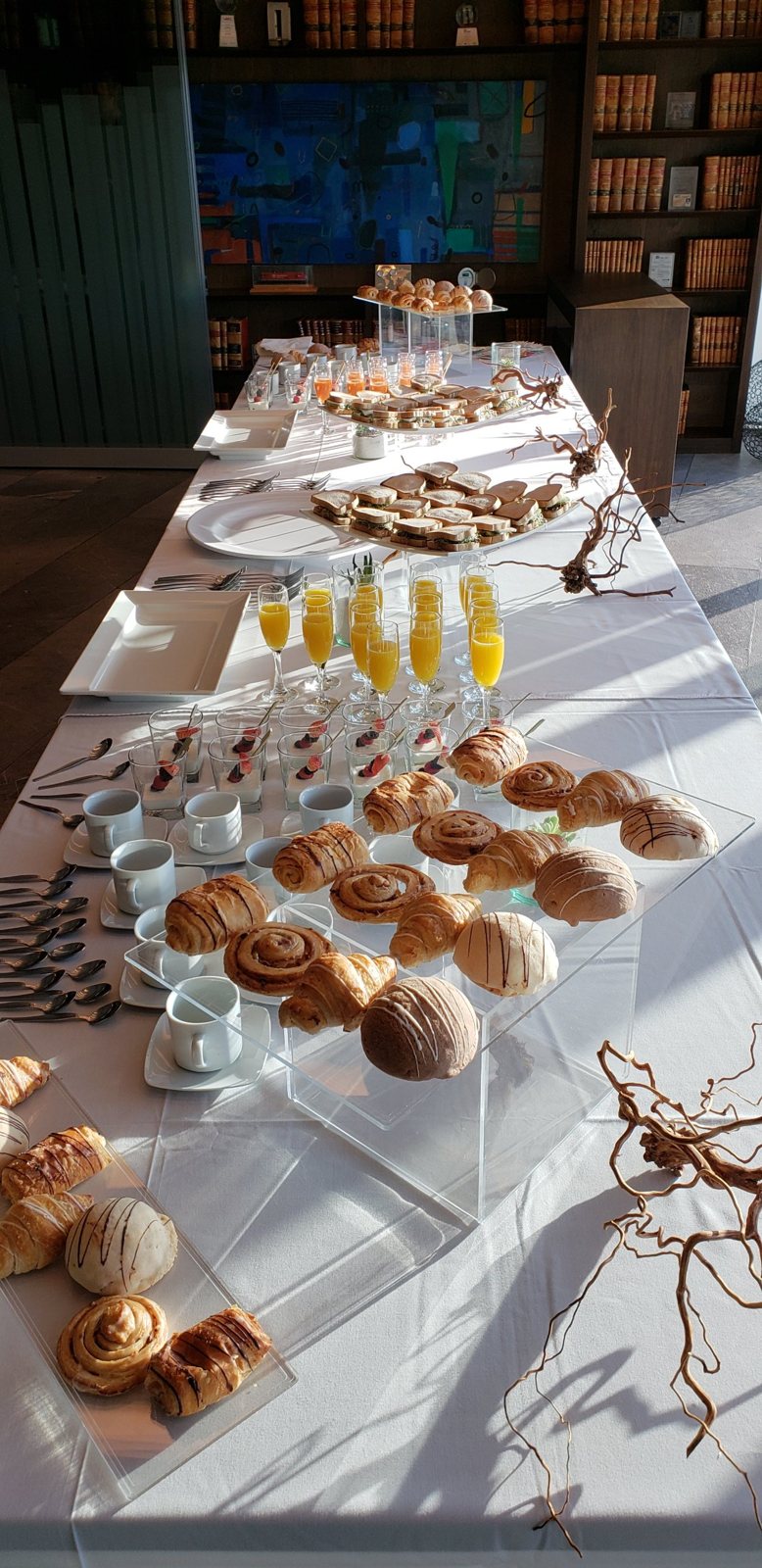 Mesa larga de desayuno empresarial con pan dulce, jugos y café en una sala con libreros